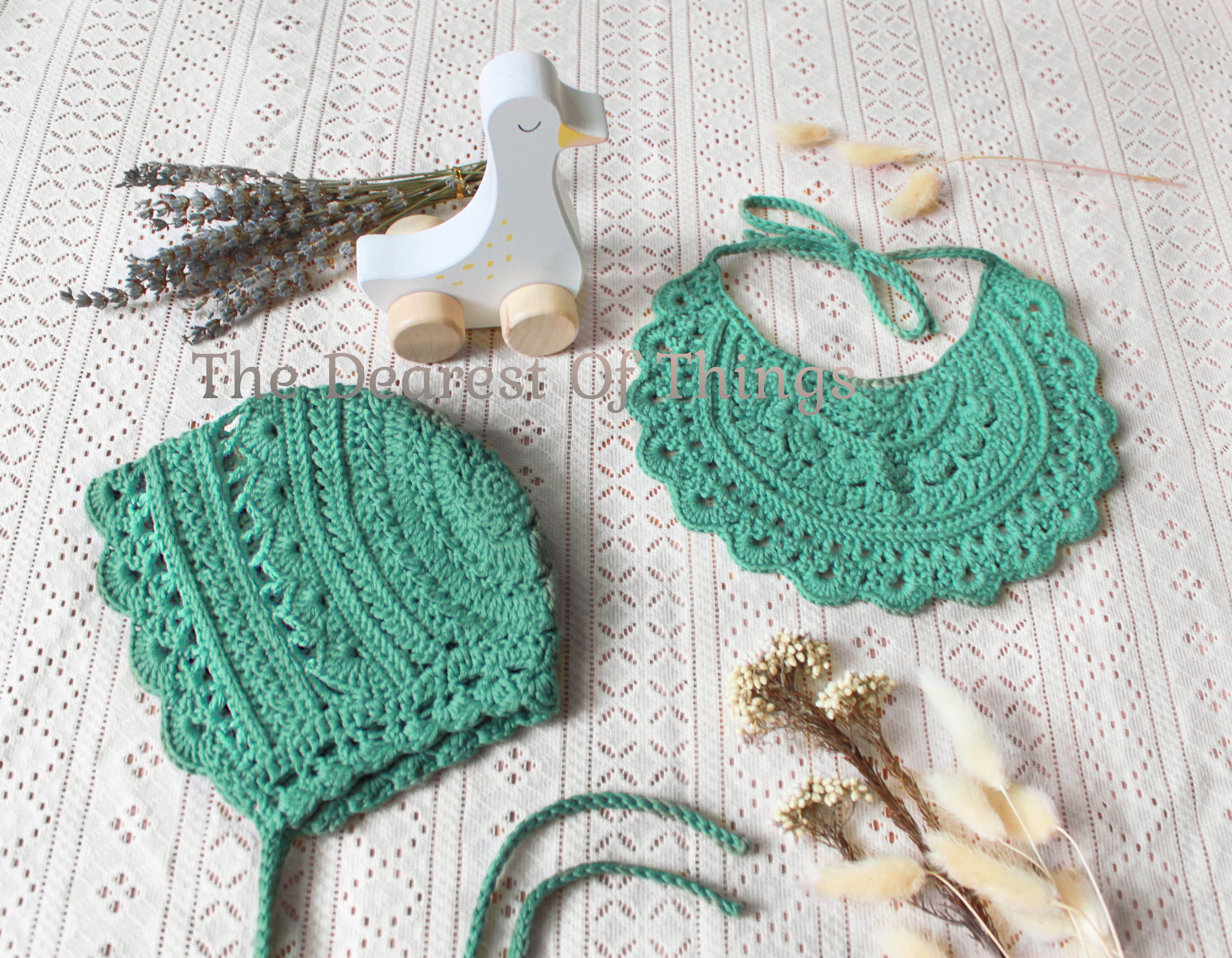 Green crocheted baby bonnet and bib on a textured surface with dried flowers.